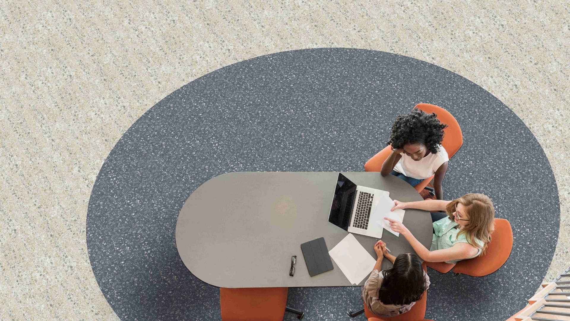 Contemporary open-plan office with patterned carpet tiles, warm wood furniture, and large windows bringing in natural light.